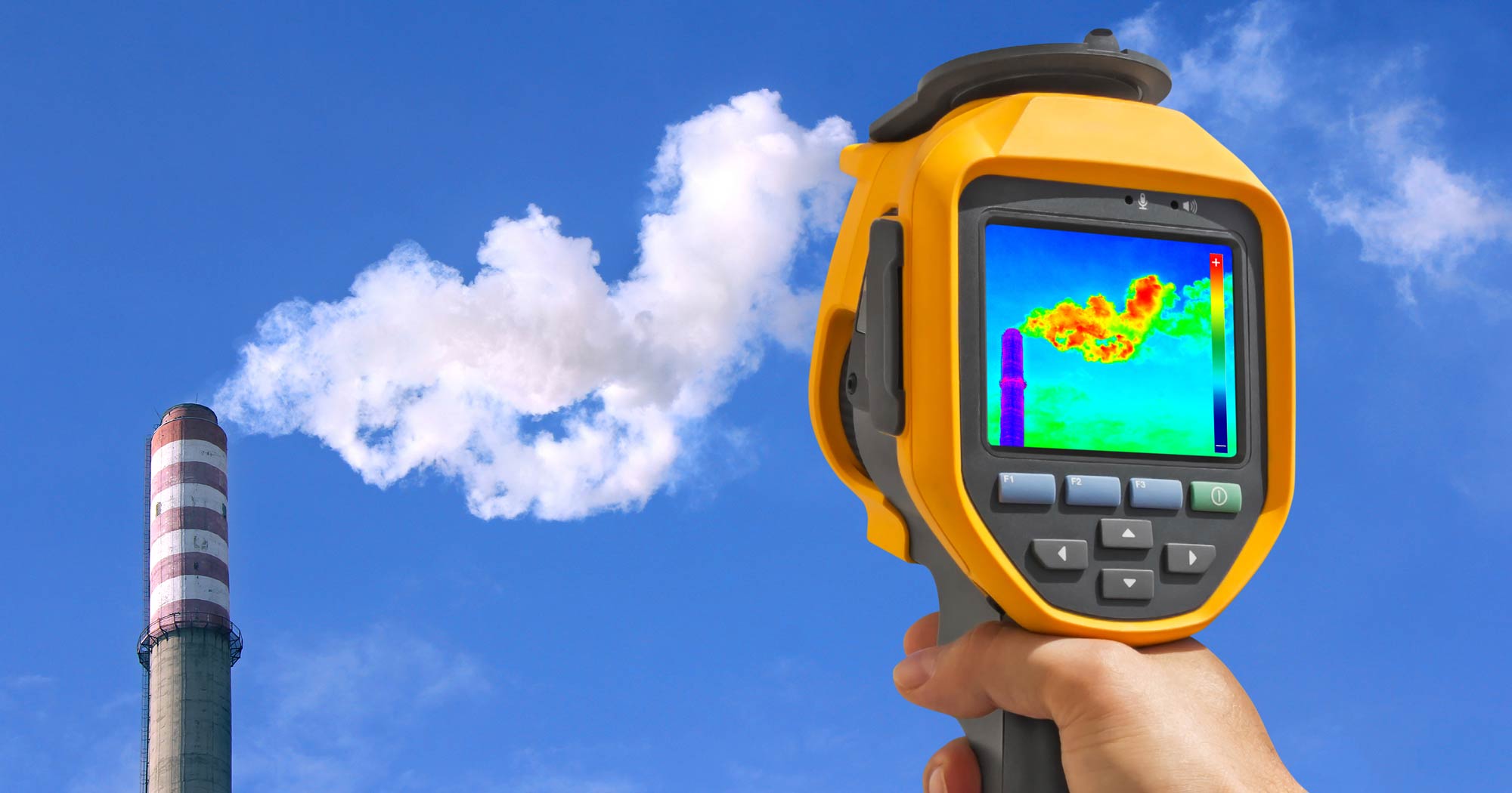 A hand holds a device recording the heat being emitted by a factory chimney.